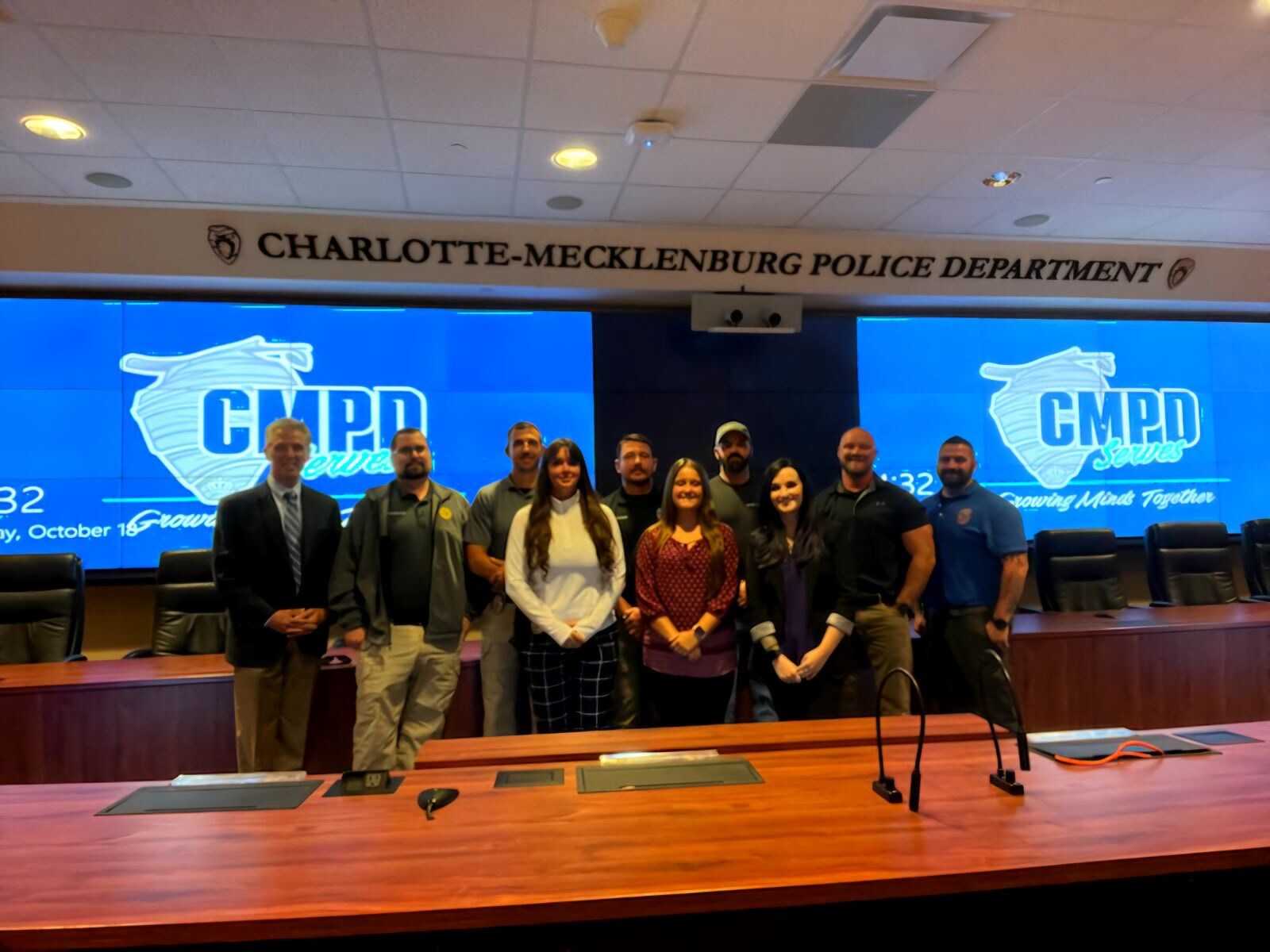 Group of ten diverse individuals posing for a photo in front of a blue backdrop with the Charlotte-Mecklenburg Police Department logo and the text 'CMPD Serves'.