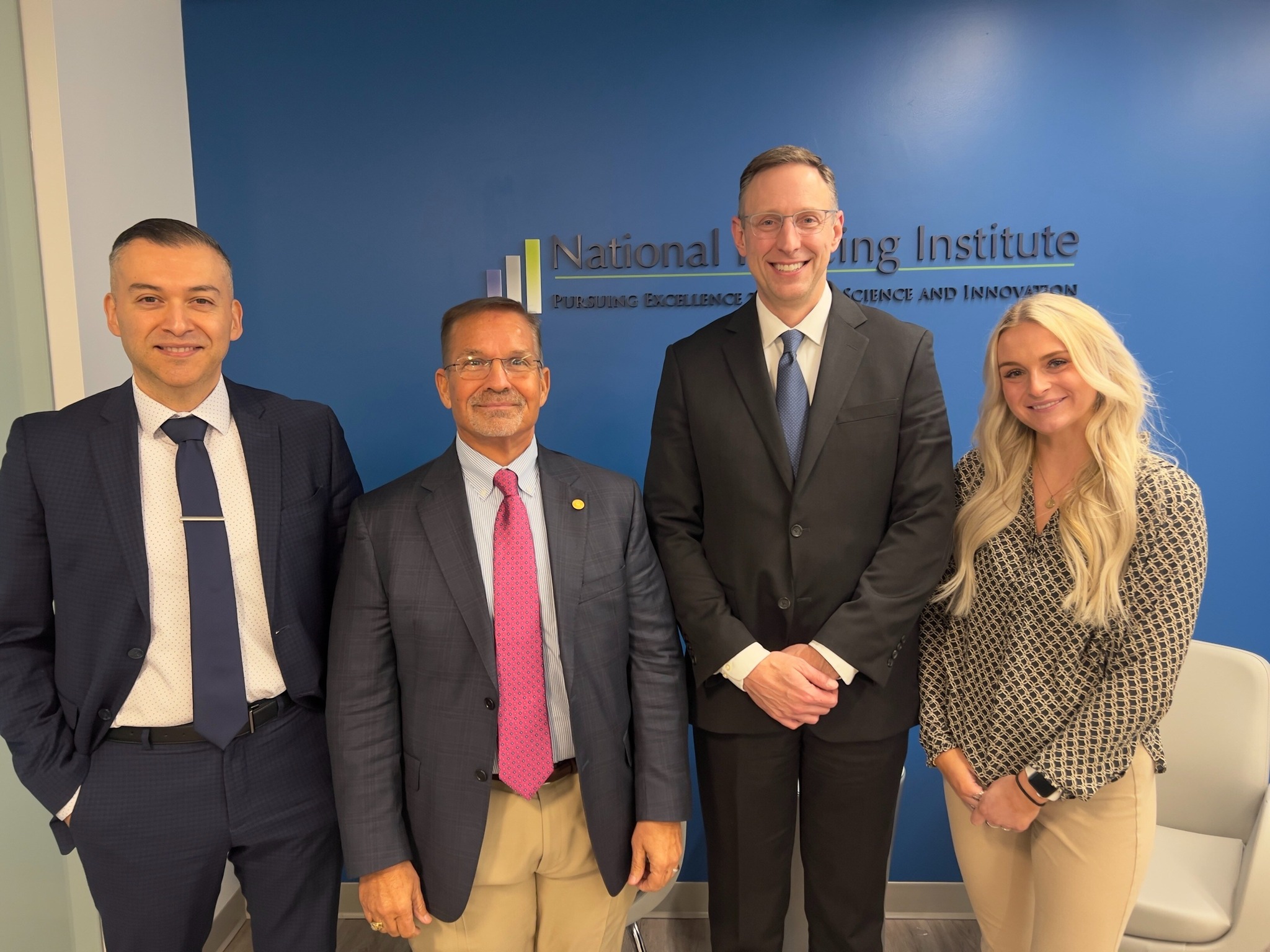 Four professional individuals, three men and one woman, standing in a line in front of a blue wall with the National Policing Institute logo. The men are dressed in business suits, while the woman is in a patterned blouse and tan trousers, all smiling at the camera.