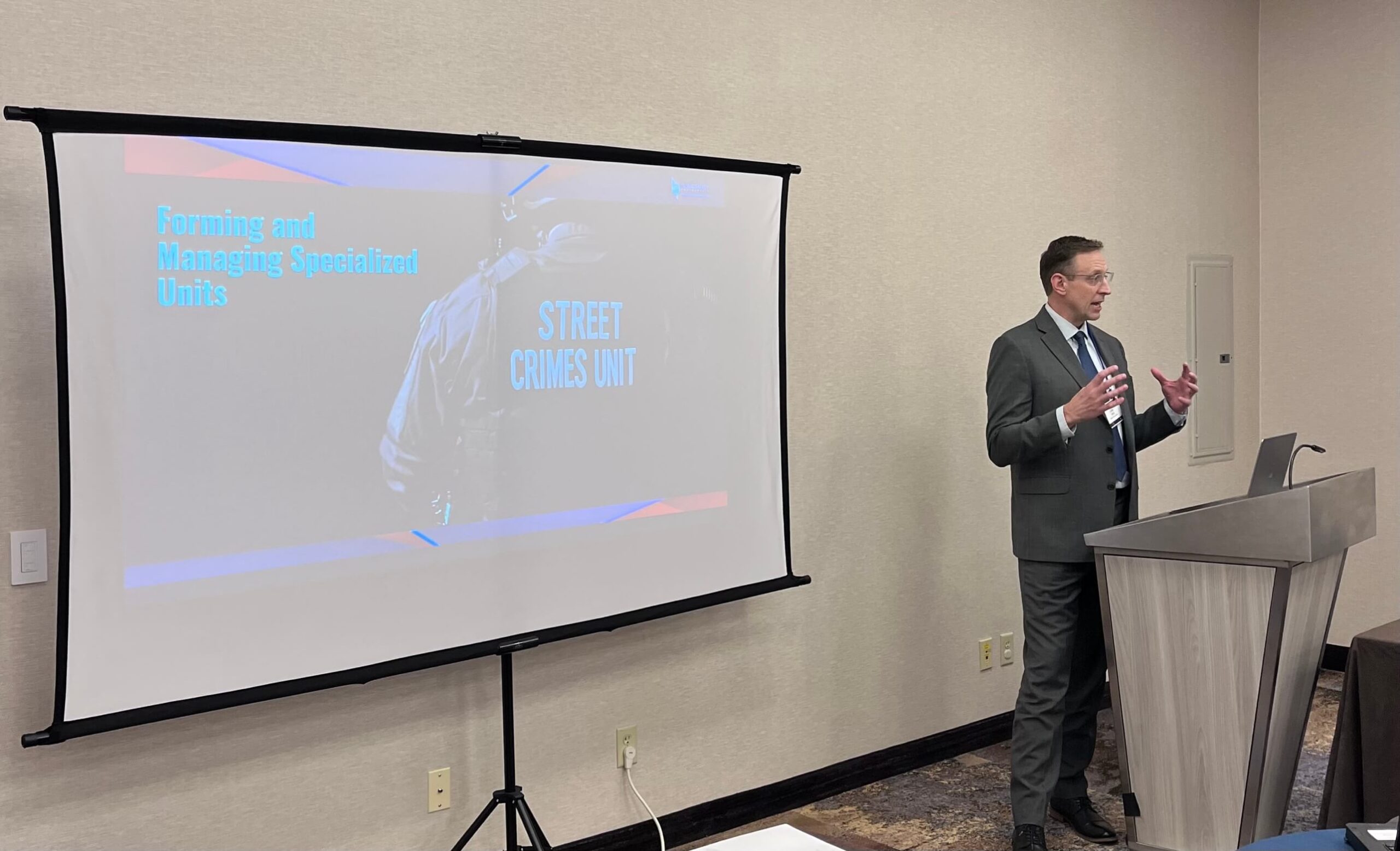 A man in a suit gestures while speaking at a podium in a conference room, with a large screen behind him displaying a presentation titled "Forming and Managing Specialized Units: Street Crimes Unit".